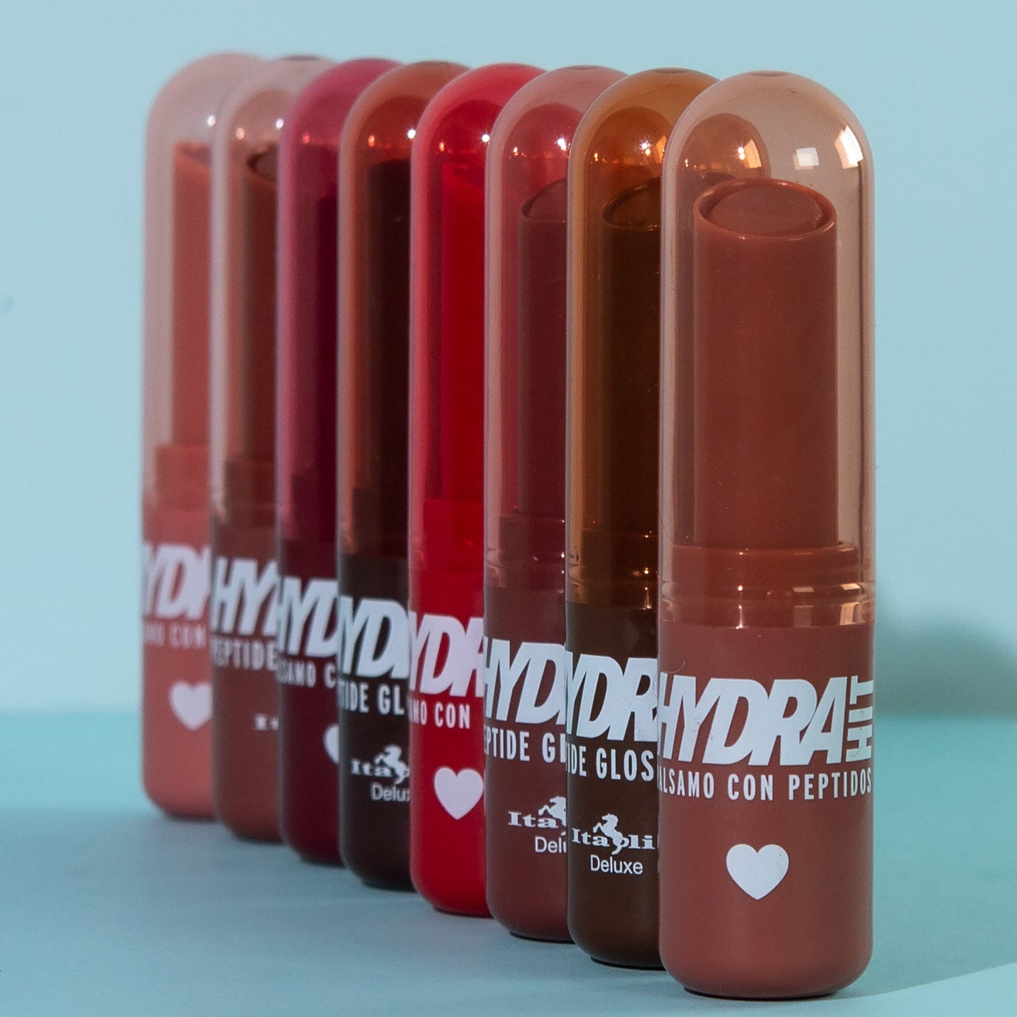 Six Hydra Hit  lip gloss balm tubes in a gradient of red, pink, and brown shades, lined up on a light blue surface against a matching light blue background.