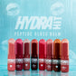 Eight Hydrahit Peptide Gloss Balm tubes in various shades of pink, red, and brown, lined up on a clear glass surface with water droplets against a textured light blue background with the brand logo.