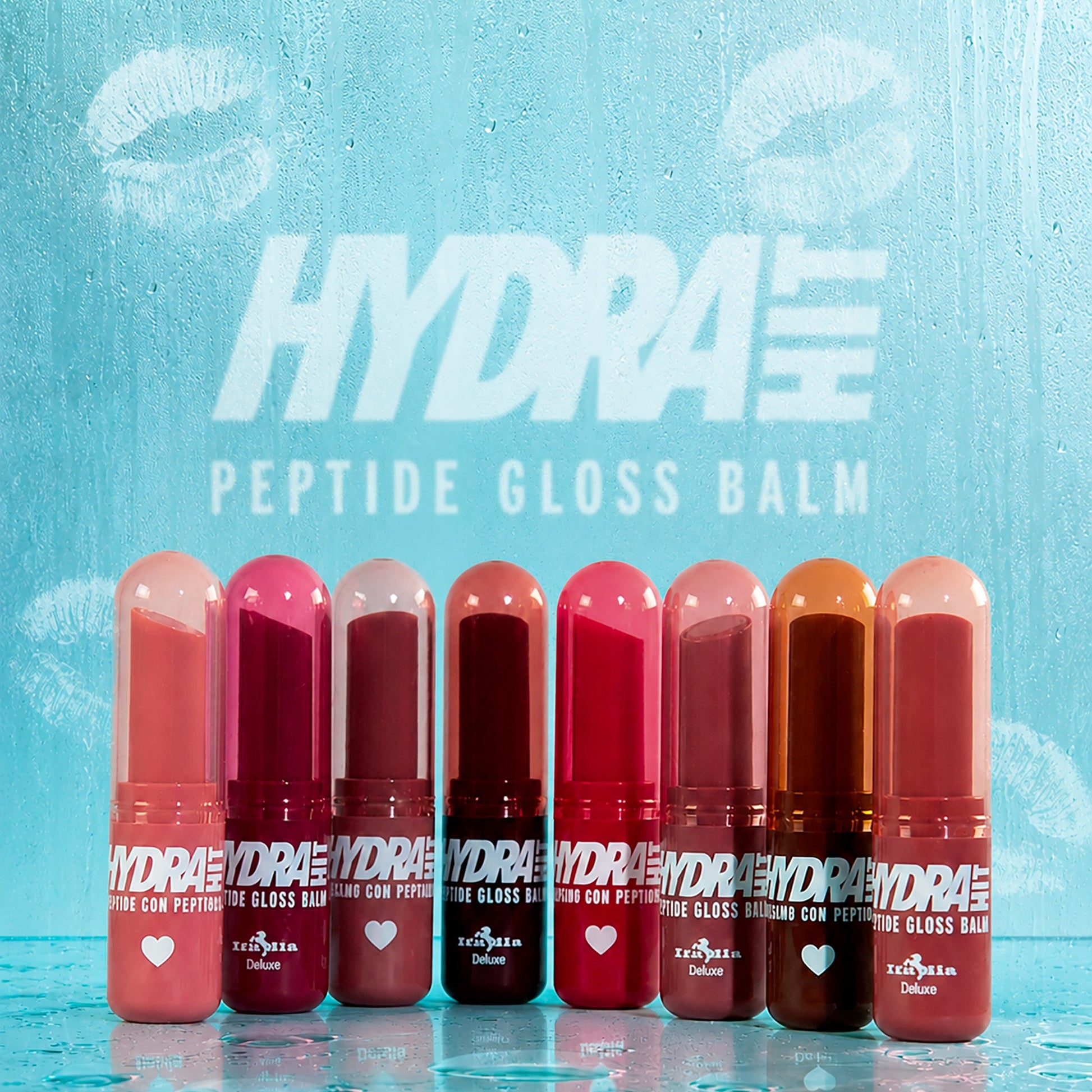 Eight Hydrahit Peptide Gloss Balm tubes in various shades of pink, red, and brown, lined up on a clear glass surface with water droplets against a textured light blue background with the brand logo.