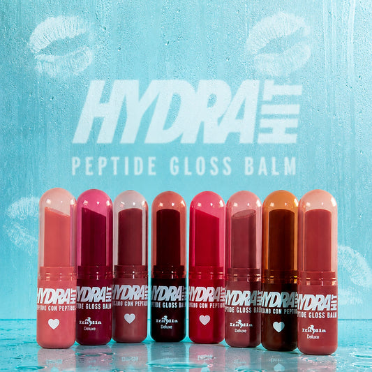 Eight Hydrahit Peptide Gloss Balm tubes in various shades of pink, red, and brown, lined up on a clear glass surface with water droplets against a textured light blue background with the brand logo.